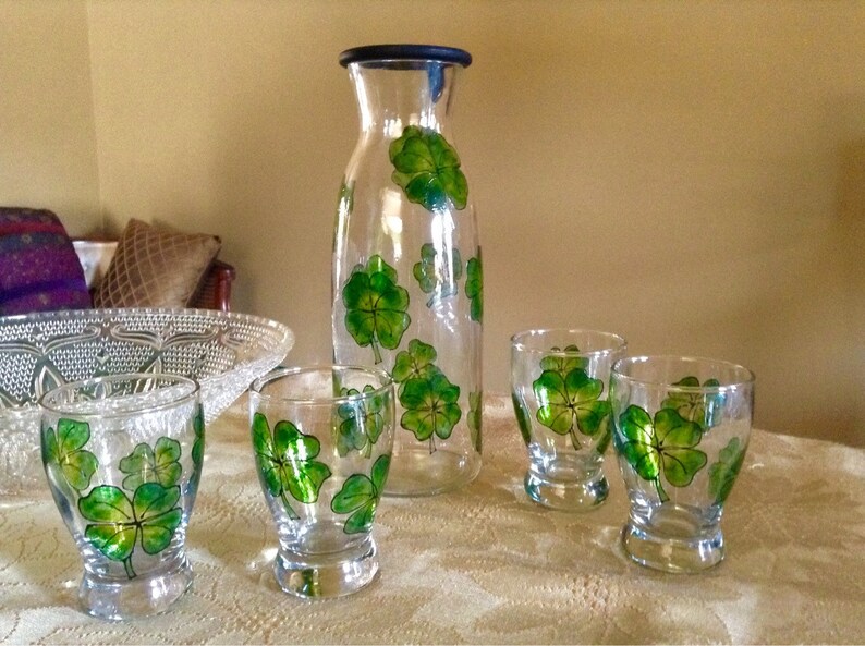 May include: A set of four clear glass drinking glasses and a matching glass pitcher, all decorated with hand-painted green four-leaf clover designs.