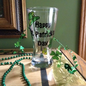 May include: A clear glass with a green shamrock design and the text "Happy St. Patrick's Day". The glass is decorated with a green garland of shamrocks.