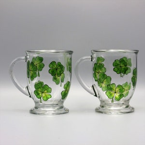 May include: Two clear glass mugs with handles, each decorated with green four-leaf clover designs.