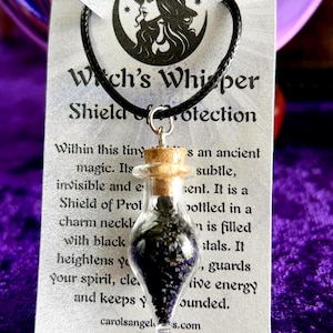 May include: A small glass vial necklace filled with dark crystals, suspended from a black cord. The tag reads "Witch's Whisper Shield of Protection" with a witch silhouette. The necklace is designed to guard and cleanse your spirit.