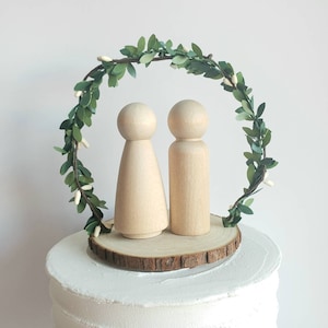 May include: A wooden cake topper featuring two small wooden figures standing under a green leafy wreath with white flowers. The figures are standing on a wooden slice.