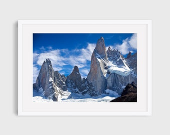 May include: A panoramic view of snow-capped mountain peaks against a bright blue sky with white clouds. The mountains are jagged and rocky, with some areas covered in snow. The sky is clear and bright, with a few fluffy clouds.
