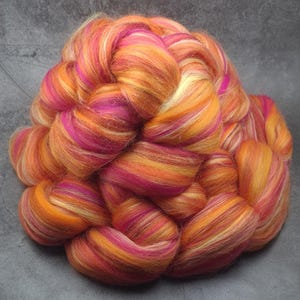 May include: A bundle of colorful wool roving in shades of orange, pink, and white. The roving is arranged in a loose braid, showing the individual fibers.