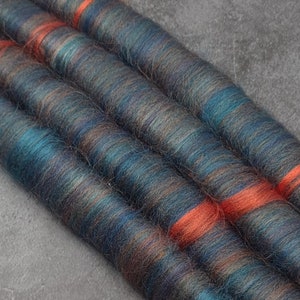 May include: Four strands of hand-spun yarn in shades of blue, teal, brown, and orange. The yarn is arranged in a row, with the orange strand running along the top of the other strands.