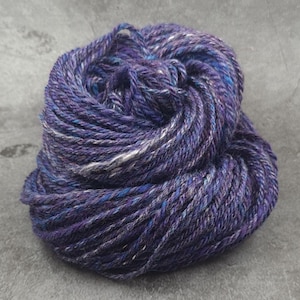 May include: A skein of hand-dyed yarn in shades of purple, blue, and white. The yarn is tightly wound, revealing the texture and color variations. Ideal for knitting, crocheting, or other fiber arts projects.