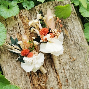 May include: Two small bouquets of dried flowers, each with a white flower, orange flowers, and dried grasses. The bouquets are tied with twine and are arranged on a piece of weathered wood.