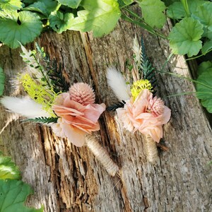 May include: Two peach-colored flower boutonnieres with dried grasses and white feathers. The boutonnieres are tied with twine and are sitting on a piece of weathered wood.