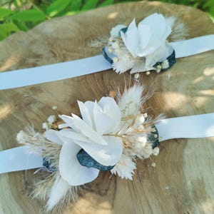 May include: Two white flower wrist corsages with dried flowers and greenery on a wooden surface. The corsages are tied with white ribbon.