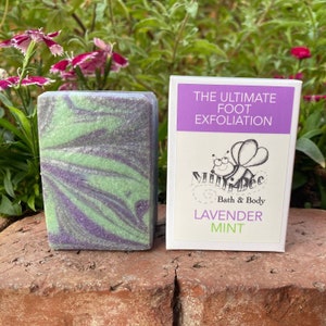 May include: A bar of lavender mint foot scrub soap with a green and purple swirl pattern. The soap is packaged in a white box with the text "The Ultimate Foot Exfoliation" and a cartoon bee with the text "Mini Bee Bath & Body" and "Lavender Mint".