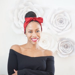 May include: A woman with a red velvet headband tied in a bow, wearing a black off-the-shoulder top, smiling at the camera.