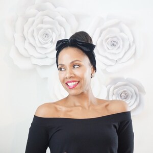 May include: A woman with dark brown skin is wearing a black velvet headband with a bow. She is smiling and looking to the right. She is wearing a black off-the-shoulder top. The background is white with large paper flowers.