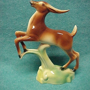 May include: A ceramic figurine of a brown deer leaping up from a green base. The deer has a long neck and a long, pointed snout.