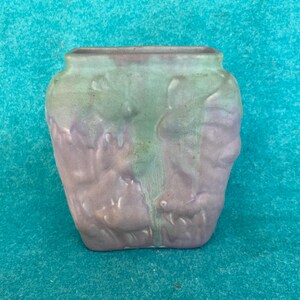 May include: A ceramic vase with a square top and a textured surface. The vase is primarily lavender with green and gray accents. The vase has a unique design and is suitable for home decor.