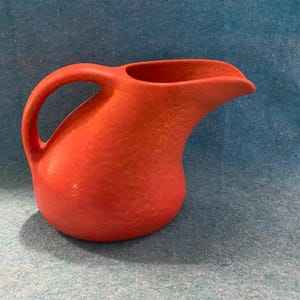 May include: A red ceramic pitcher with a textured surface and a curved spout. The pitcher has a handle and a wide opening.
