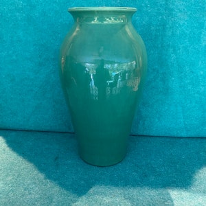 May include: A tall, teal-colored ceramic vase with a glossy finish. The vase has a wide body that tapers towards the base and a slightly flared rim. The background is a textured teal surface.