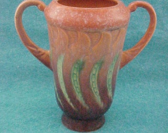 May include: A brown ceramic creamer with green and brown floral designs. The creamer has two handles and a rounded body.