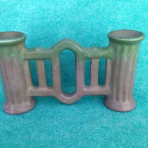 May include: A green and purple ceramic vase with a decorative fence design. The vase has two openings for flowers.