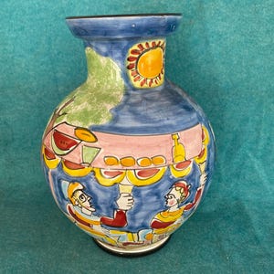 May include: A hand-painted ceramic vase with a blue background and a scene of two figures under a yellow umbrella. The figures are wearing colorful clothing and holding glasses. The vase has a round body and a narrow neck.