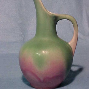 May include: A green and pink ceramic pitcher with a wide, rounded body and a curved handle. The pitcher has a glazed finish with a subtle ombre effect.