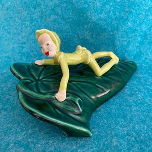 May include: A ceramic figurine of a green elf laying on a large green leaf. The elf is wearing a yellow outfit and has a red mouth.