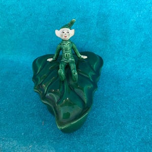 May include: A green ceramic leaf with a small green and white elf figurine sitting on top. The elf is wearing a green hat and a green outfit.