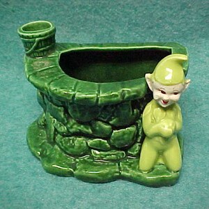 May include: A green ceramic planter shaped like a well with a small bucket on top. A green and white elf figurine is standing next to the well.
