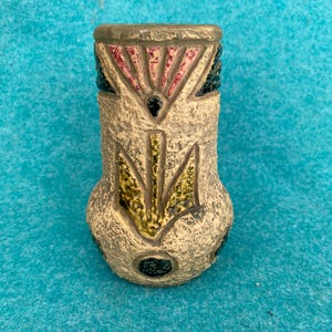 May include: A textured, beige ceramic vase with a flared top and a rounded base. The vase features a geometric design with red, green, and black accents. The top rim is a darker shade of brown. The vase is approximately 4 inches tall.