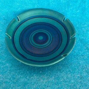 May include: A round ceramic ashtray featuring a series of concentric circles in shades of teal, turquoise, and deep navy blue. The rim of the ashtray includes four evenly spaced notches designed for resting items, with a smooth, glazed finish.