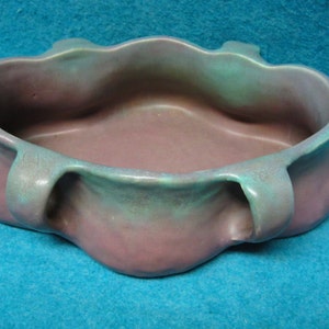 May include: A purple and green ceramic bowl with a wavy rim and three handles. The bowl is glazed with a mottled finish.