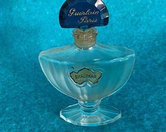 Könnte beinhalten: Eine durchsichtige Glasflasche mit Shalimar-Parfum von Guerlain Paris. Die Flasche hat ein dekoratives, gewelltes Design und einen blauen, facettierten Verschluss mit dem Markennamen.