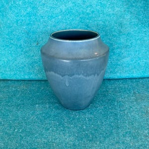 May include: A blue ceramic vase with a wide mouth and a rounded body. The vase has a matte finish and a slightly textured surface. The background is a solid teal color.
