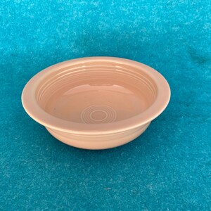 May include: A light peach-colored ceramic bowl with a wide rim and concentric rings inside. The bowl is round and sits on a blue surface. The bowl is likely for serving food.
