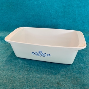 May include: A rectangular white ceramic baking dish with rounded corners and integrated handles on the short sides. The exterior features a centered blue floral pattern consisting of small flowers and leaves on the front face of the vessel.