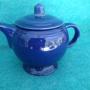 May include: A cobalt blue ceramic teapot with a lid and a round body. The teapot has a handle and a spout. The teapot is sitting on a blue surface.