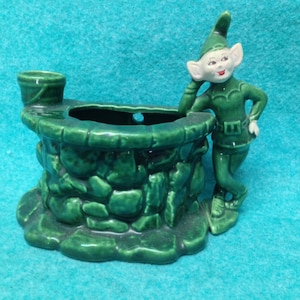 May include: A green ceramic planter shaped like a stone well with a small bucket on top. A green elf figurine leans against the well, with a red nose and white eyes.