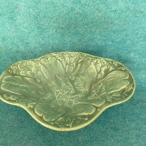 May include: A light green, leaf-shaped ceramic dish with a detailed embossed design. The dish has a scalloped edge and a matte finish. The design features a botanical pattern of leaves and stems.