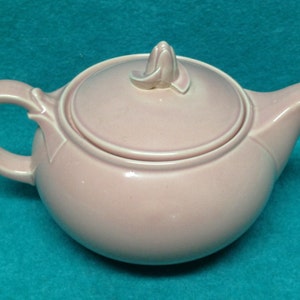 May include: A pink ceramic teapot with a lid and a floral design on the top. The teapot has a rounded body and a spout on the right side.