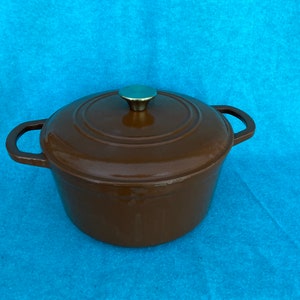 May include: A brown cast iron Dutch oven with a lid and two side handles. The lid has a gold-colored knob. The pot is on a blue background. This cookware is ideal for slow cooking and baking.