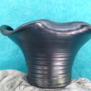 May include: A dark gray ceramic vase with a flared, ridged body and a wavy, rolled rim. The vase sits on a light-colored cloth against a turquoise background. The vase has a smooth, metallic finish.