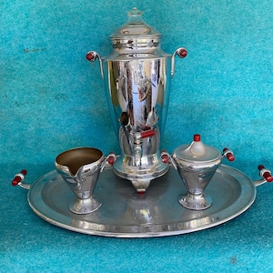 May include: A silver coffee percolator with red handles, a creamer, a sugar bowl, and a silver tray. The set is on a blue background.