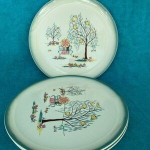 May include: Three white ceramic plates with a green rim. Each plate features a whimsical illustration of a couple sitting on a bench under a tree with yellow flowers. The trees are depicted with blue and green leaves and the background is a light blue.