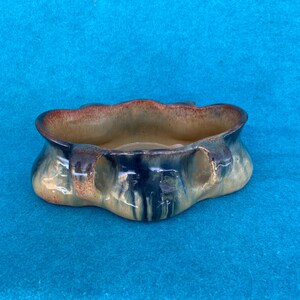 May include: A ceramic bowl with a unique, wavy rim and a multi-toned glaze. The bowl features a blend of colors, including brown, blue, and tan. The exterior has a textured appearance, and the interior is smooth. The bowl is set against a blue background.