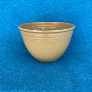 May include: A light yellow ceramic bowl with a rounded shape and a slightly flared rim. The bowl has a smooth exterior with two horizontal ridges near the top. The interior is a slightly darker shade of yellow. The bowl is set against a blue background.