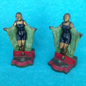 May include: A pair of vintage bookends featuring a woman in a black swimsuit and a green shawl. The bookends are made of metal and have a gold finish. The bases are red and gold.