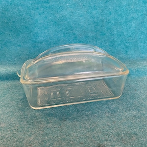May include: A clear glass butter dish with a rectangular base and a matching arched lid. The base features a textured, embossed pattern on the bottom surface, while the lid has a smooth, curved top and a small handle tab on one side.