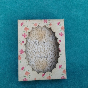 May include: A vintage perfume box with a white background and pink floral accents. The box has a scalloped edge and a cutout in the center that says "Lilas Perfume".