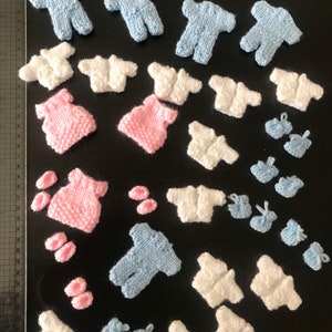 May include: A collection of miniature knitted baby clothes in blue, pink, and white. The clothes include onesies, sweaters, and booties.