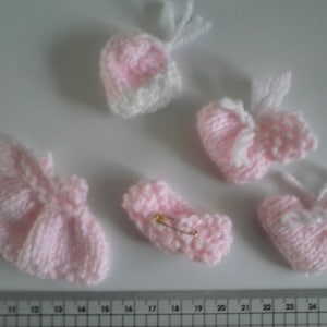Könnte beinhalten: Ein Set an rosa gestrickten Babykleidern, bestehend aus einer Mütze, Stiefeln und einem Kleid. Die Artikel sind alle aus einem weichen, zarten Garn gefertigt und perfekt für ein Neugeborenes.