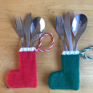 Puede incluir: Dos calcetines de Navidad de punto en rojo y verde con ribetes blancos, cada uno con un tenedor, un cuchillo y una cuchara.