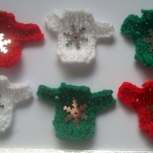 Könnte beinhalten: Sechs kleine gestrickte Weihnachtspullover mit Schneeflocken-Verzierungen. Die Pullover sind rot, weiß und grün.